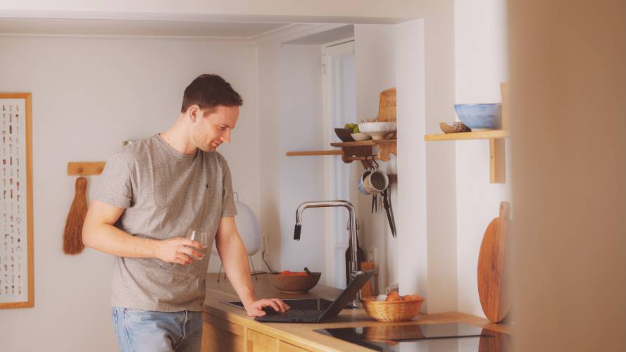 Ung mann smiler med laptop og vannglass ved kjøkkenbenken