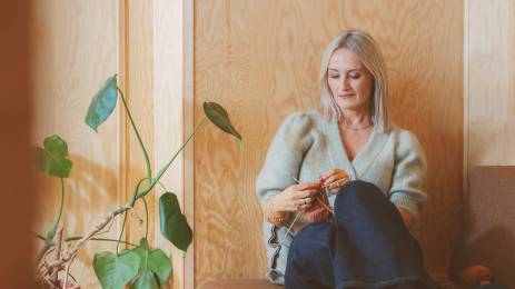 Voksen kvinne i sofa ser ned på strikketøyet sitt