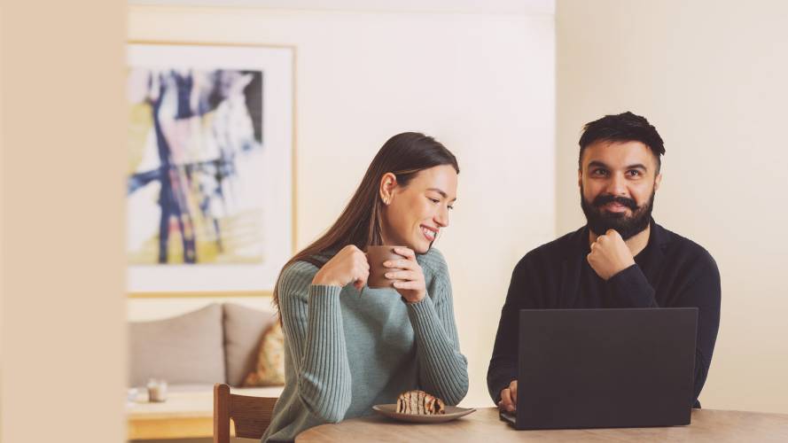 Ungt smilende par sitter ved kjøkkenbordet med kaffekopp og laptop
