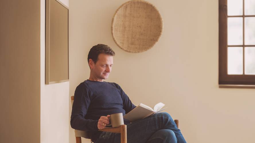 Voksen mann leser bok i stol med kaffekopp i hånd