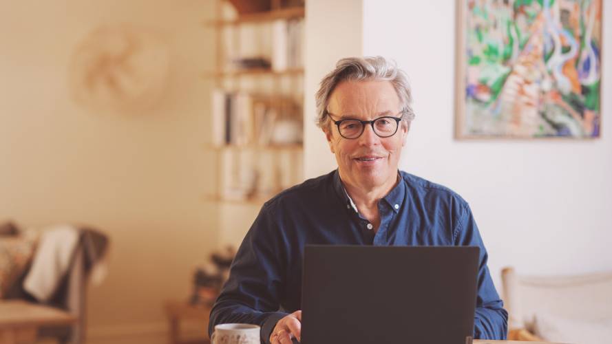 Voksen mann sitter ved kjøkkenbord med laptop og kaffekopp