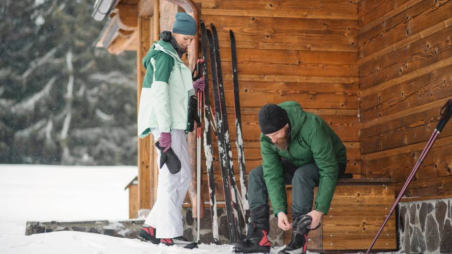 Mann og dame som tar på seg skiutstyr utenfor hytte i skogen