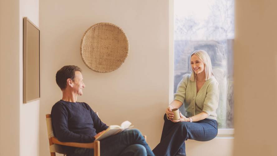 Smilende voksen mann i stol ser mot smilende kvinne i vinduskarm med kaffekopp